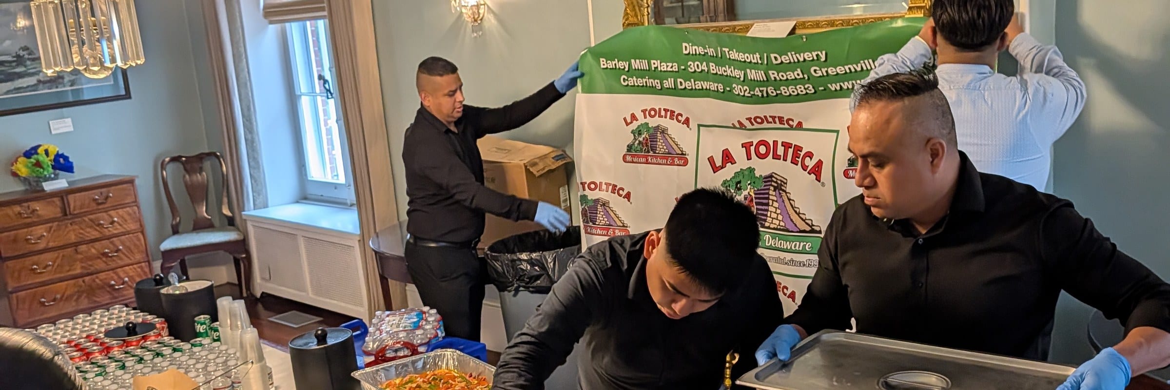 La Tolteca staff setting up a catered event with the La Tolteca banner and chafing dishes